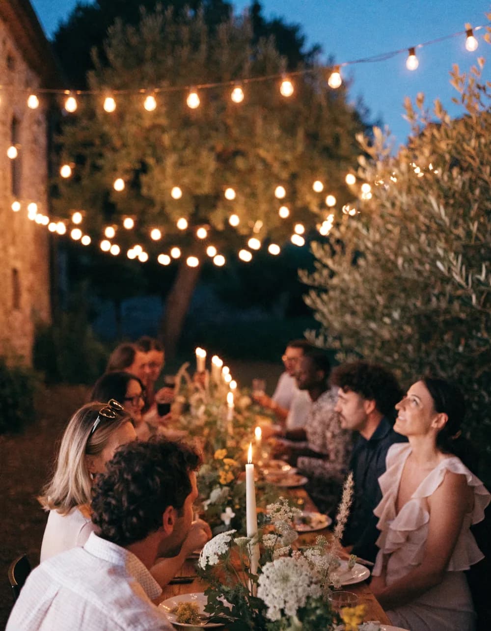 A festoon-lit communal dinner table in a villa garden at dusk.
